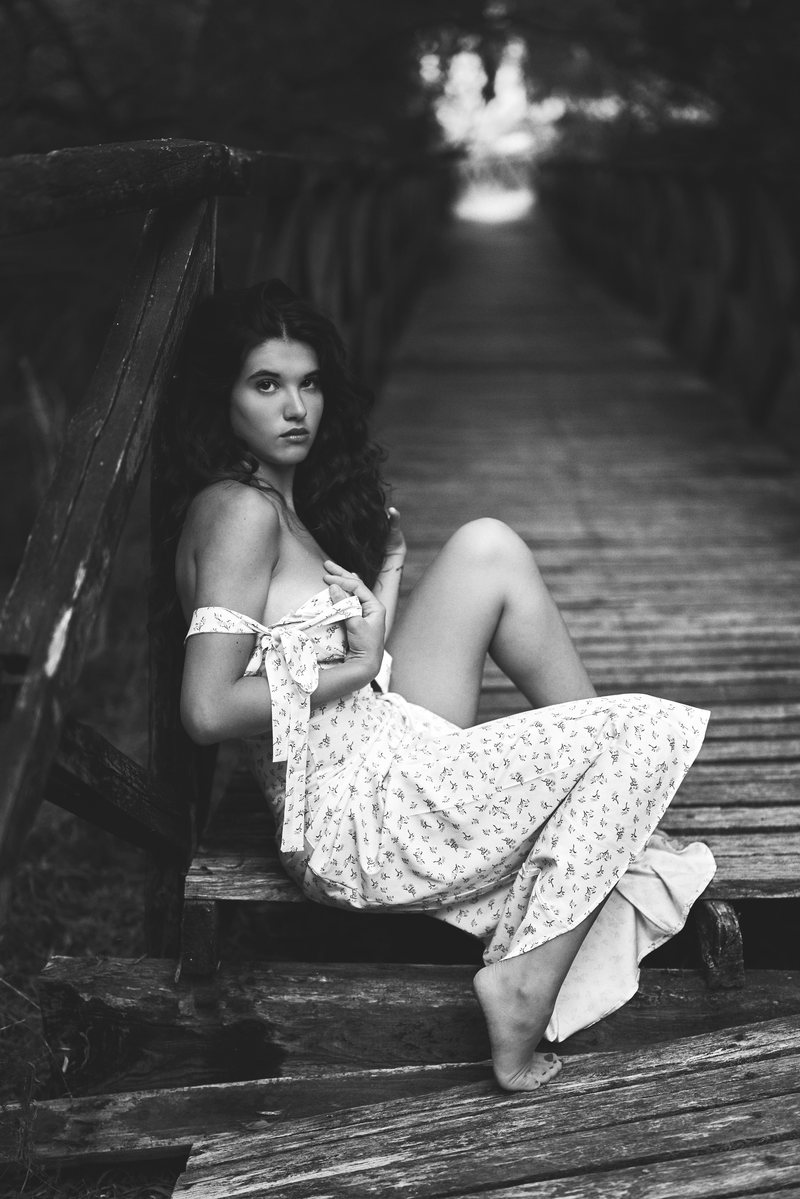 Black and white portrait of a woman in a floral dress sitting on a wooden bridge in the woods