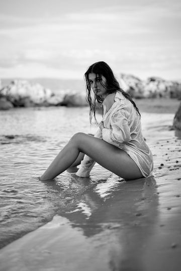 Black and white portrait of a woman in a white shirt sitting at the water's edge, her feet in the sea