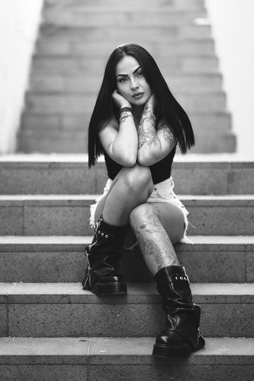 Black and white portrait of a tattooed woman sitting on a staircase in boots, her hands on her knees