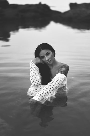 Black and white portrait of a woman in white trousers sitting in the water, her hand on her face
