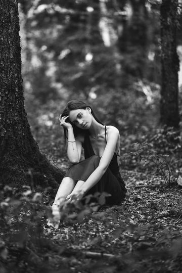 Black and white portrait of a woman sitting at the foot of a tree in the woods, her eyes lowered