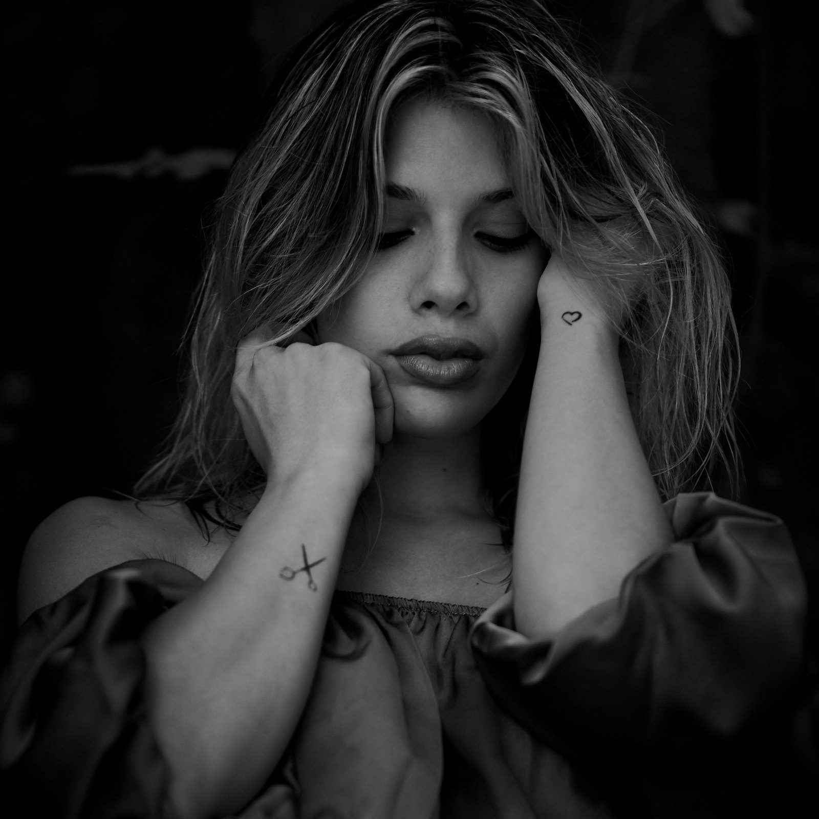 Black and white portrait of a young woman with closed eyes, her face between her hands, a small heart tattooed on her wrist, in the dark