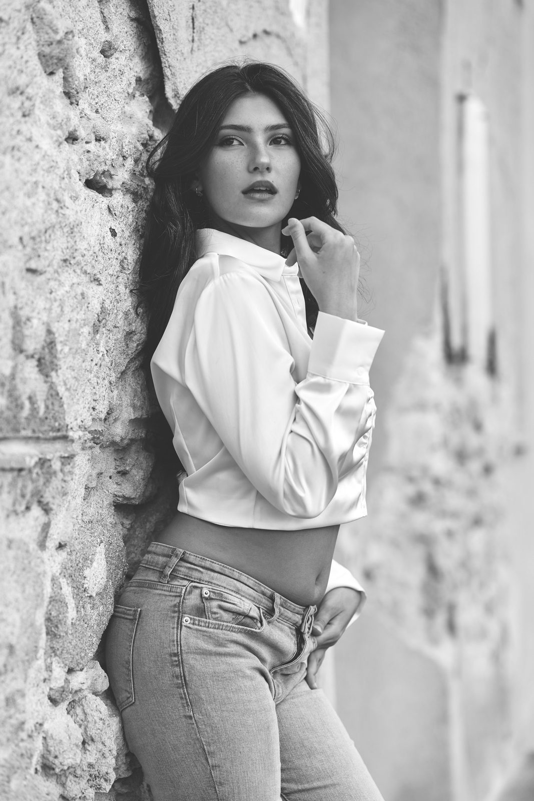 Ritratto in bianco e nero di una donna in camicia bianca appoggiata alle mura di Alghero, la mano tra i capelli