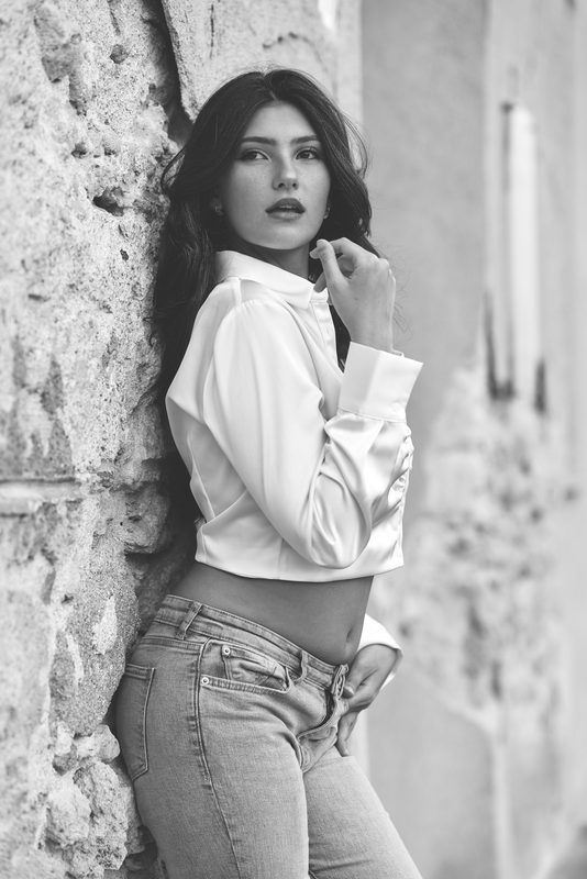 Ritratto in bianco e nero di una donna in camicia bianca appoggiata alle mura di Alghero, la mano tra i capelli