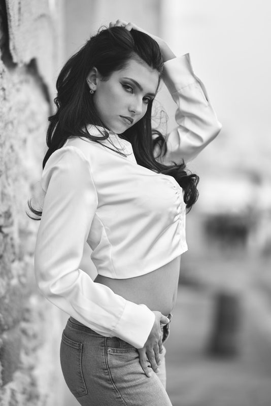Ritratto in bianco e nero di una donna in camicia bianca appoggiata a una parete di pietra, luce del mattino