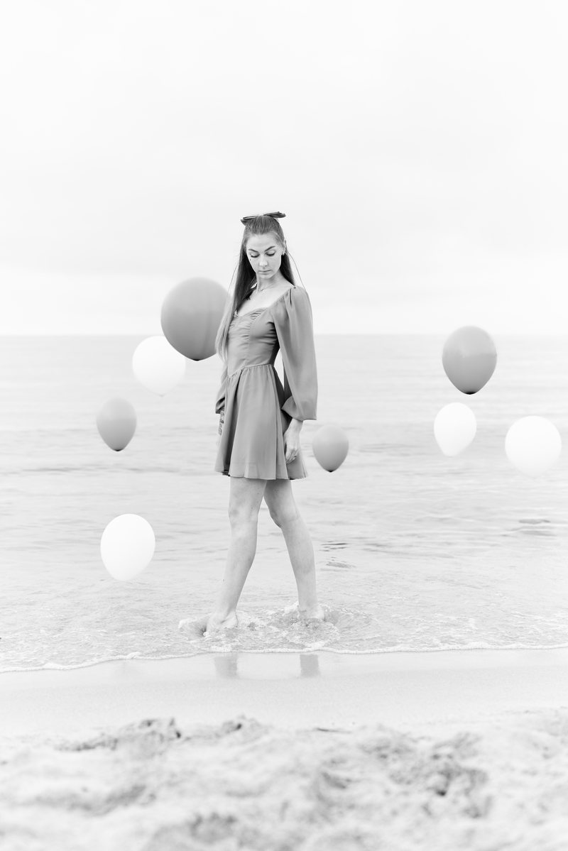 Ritratto in bianco e nero di una donna in piedi in riva al mare con dei palloncini, in abito chiaro