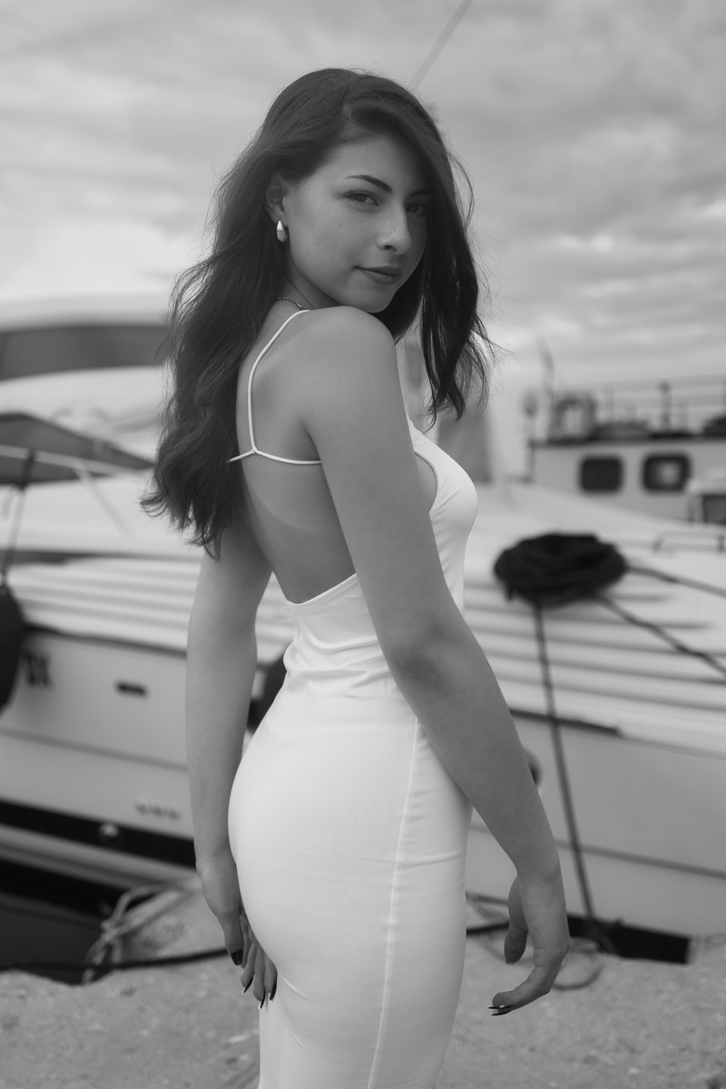 Ritratto in bianco e nero di una donna in abito bianco, di spalle accanto a una barca nel porto, con lo sguardo verso l'obiettivo