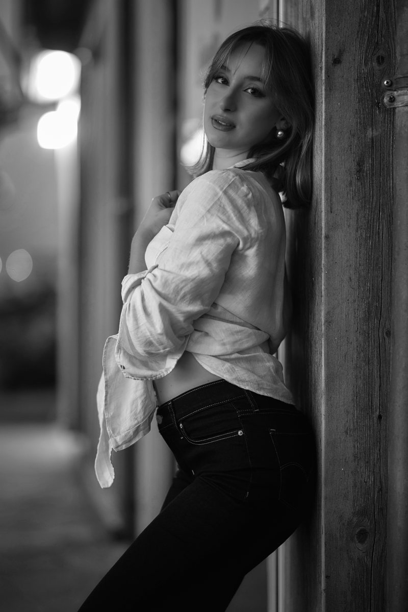 Ritratto in bianco e nero di una donna in camicia bianca appoggiata a una porta, di notte, con le luci della città dietro