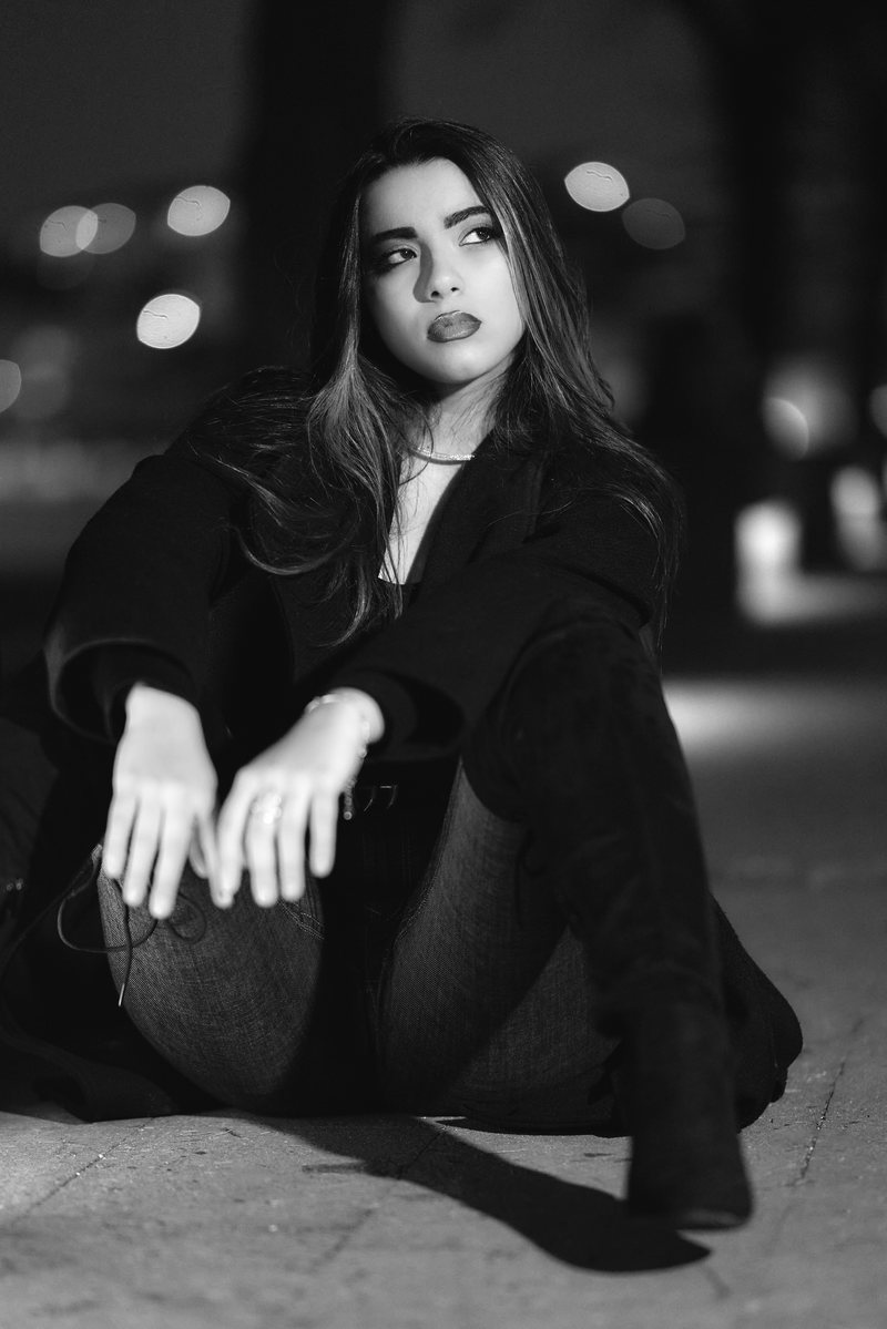 Ritratto in bianco e nero di una donna in cappotto scuro seduta a terra di notte, le mani sulle ginocchia