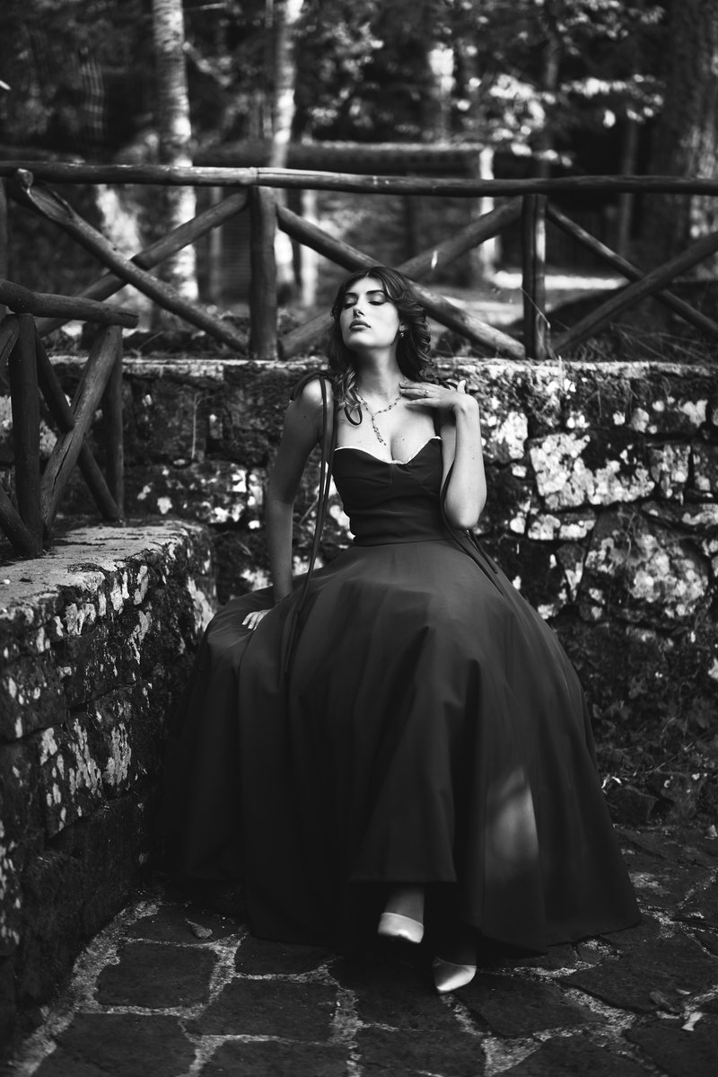 Black and white portrait of a woman in a long black dress sitting on a stone wall in the woods, eyes closed