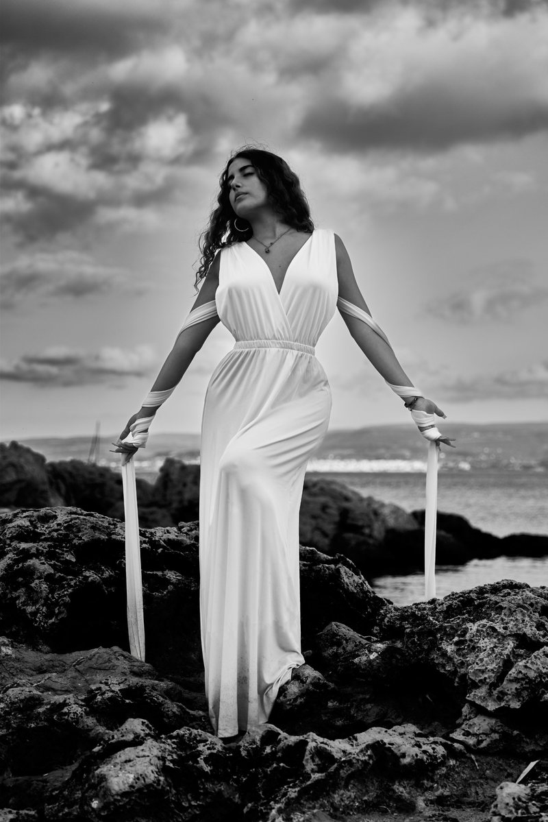 Black and white portrait of a woman in a white dress among the rocks, eyes closed, her face turned to a cloud-heavy sky