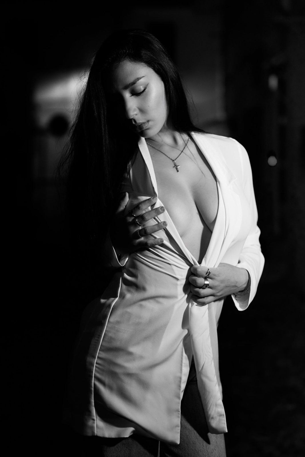 Black and white portrait of a young woman with long dark hair, her eyes closed and her face lowered, in an open white jacket held with her hands, against a dark background
