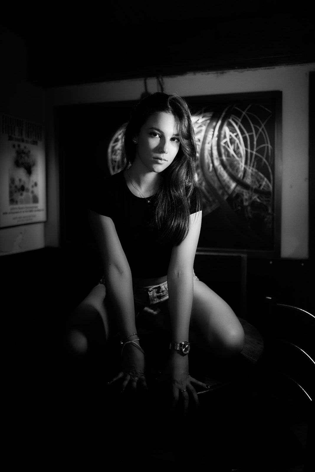 Black and white portrait of a young woman in a dark t-shirt, crouching in a dim room with her hands resting in front of her, looking straight into the camera, in front of a large abstract painting