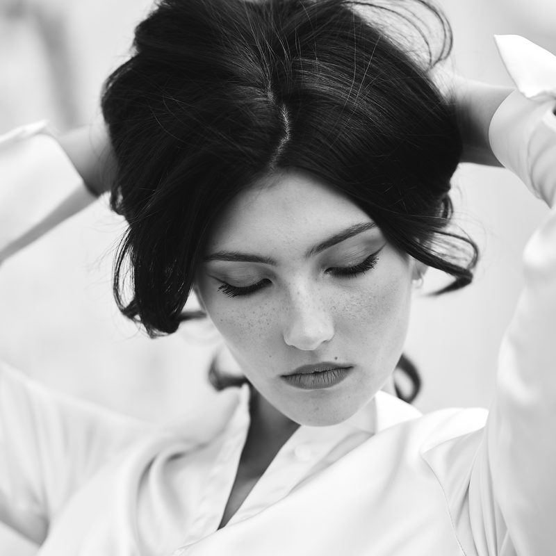 Ritratto in bianco e nero di una donna in camicia bianca, le braccia sollevate dietro la testa, sguardo abbassato